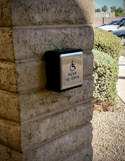 A push to open button is mounted into a brick pillar with a view of bushes and cars in a parking lot in the background.