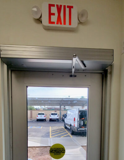 A business door with a view of a parking lot and parked vehicles. An exit sign hangs above the door.